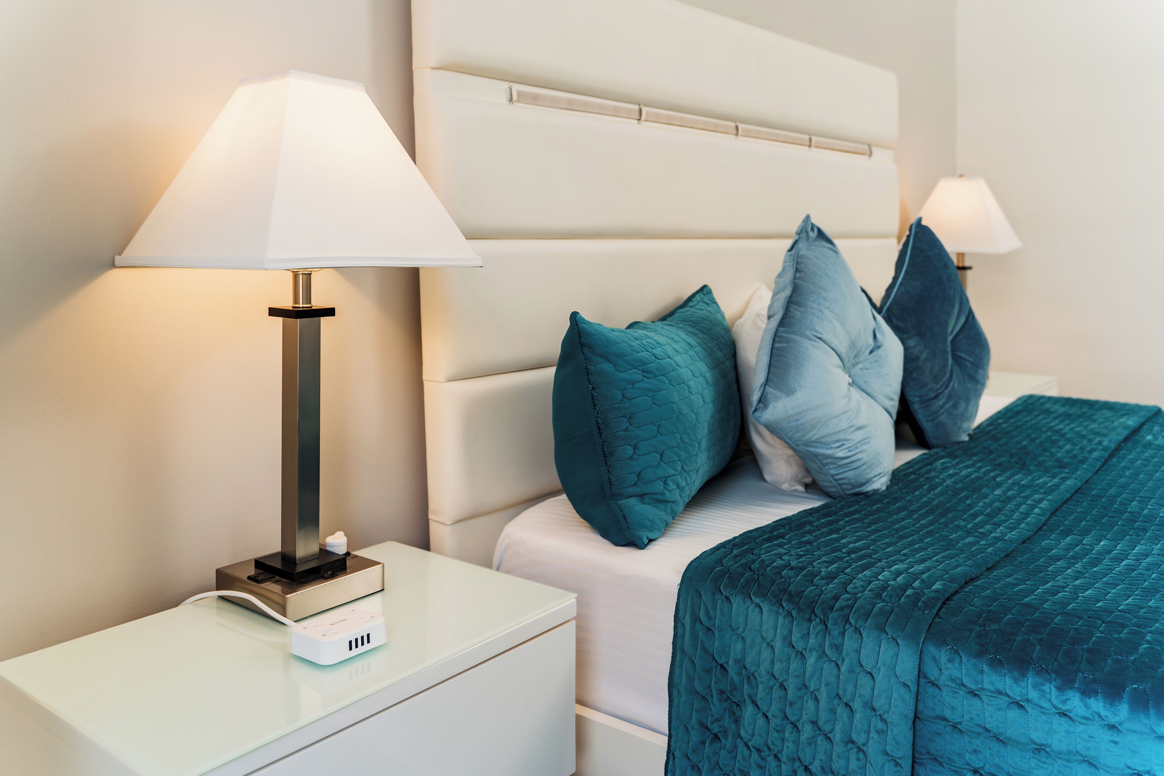 Un lit bien fait, agrémenté d’oreillers et d’une couverture de couleur turquoise, une table de chevet blanche avec une lampe, et une petite table de nuit blanche.