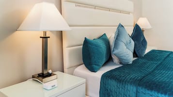 Ein ordentlich bezogenes Bett mit türkisfarbenen Kissen und einer türkisfarbenen Decke, ein weißer Nachttisch mit einer Lampe und ein weißer Beistelltisch.