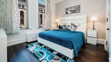 Un dormitorio moderno con una cama grande, dos mesitas de noche, una ventana con cortinas, un radiador y vistas a un edificio.