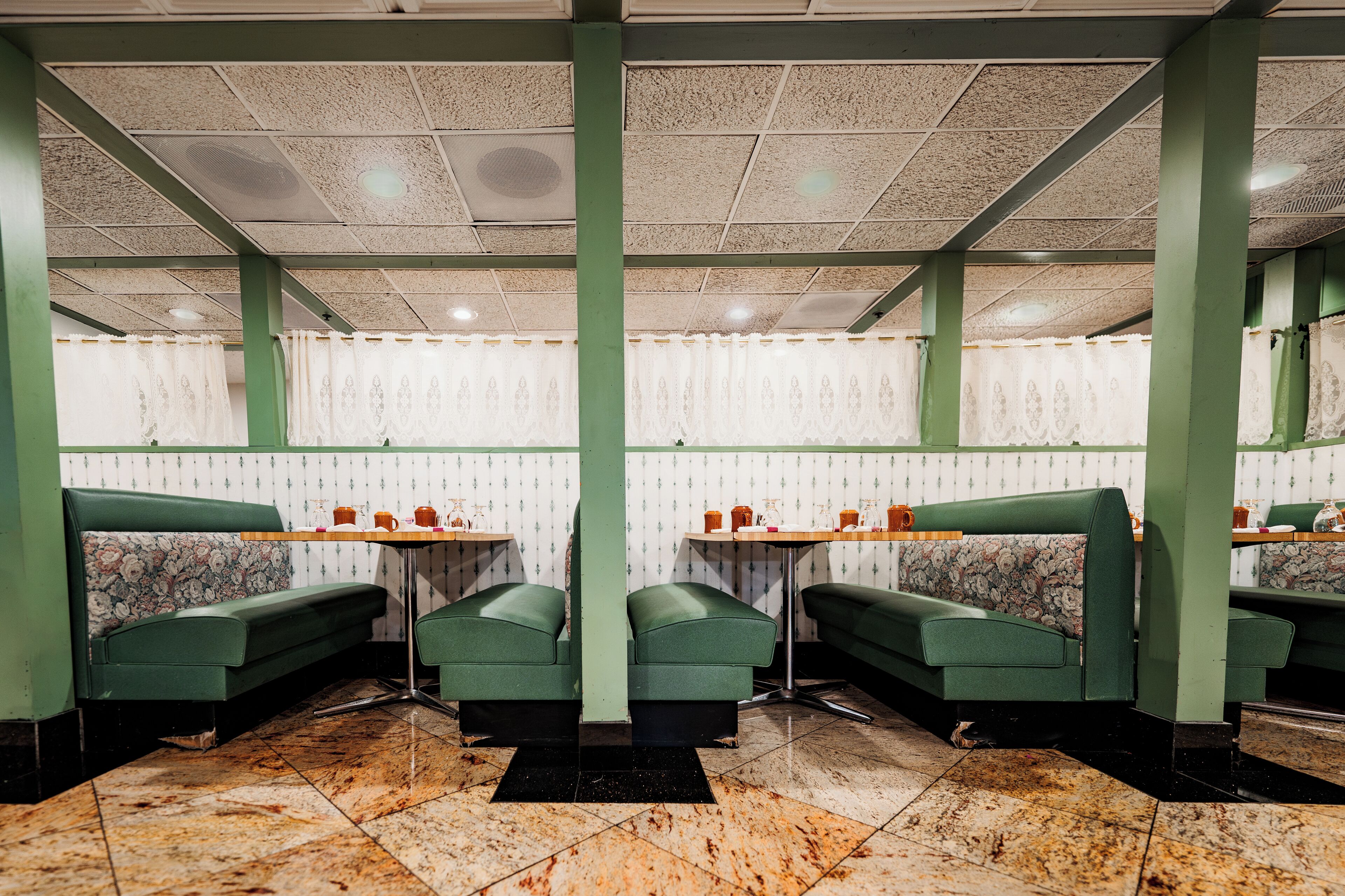 Interior de restaurante com bancos verdes, piso de mármore, papel de parede estampado e iluminação no teto.