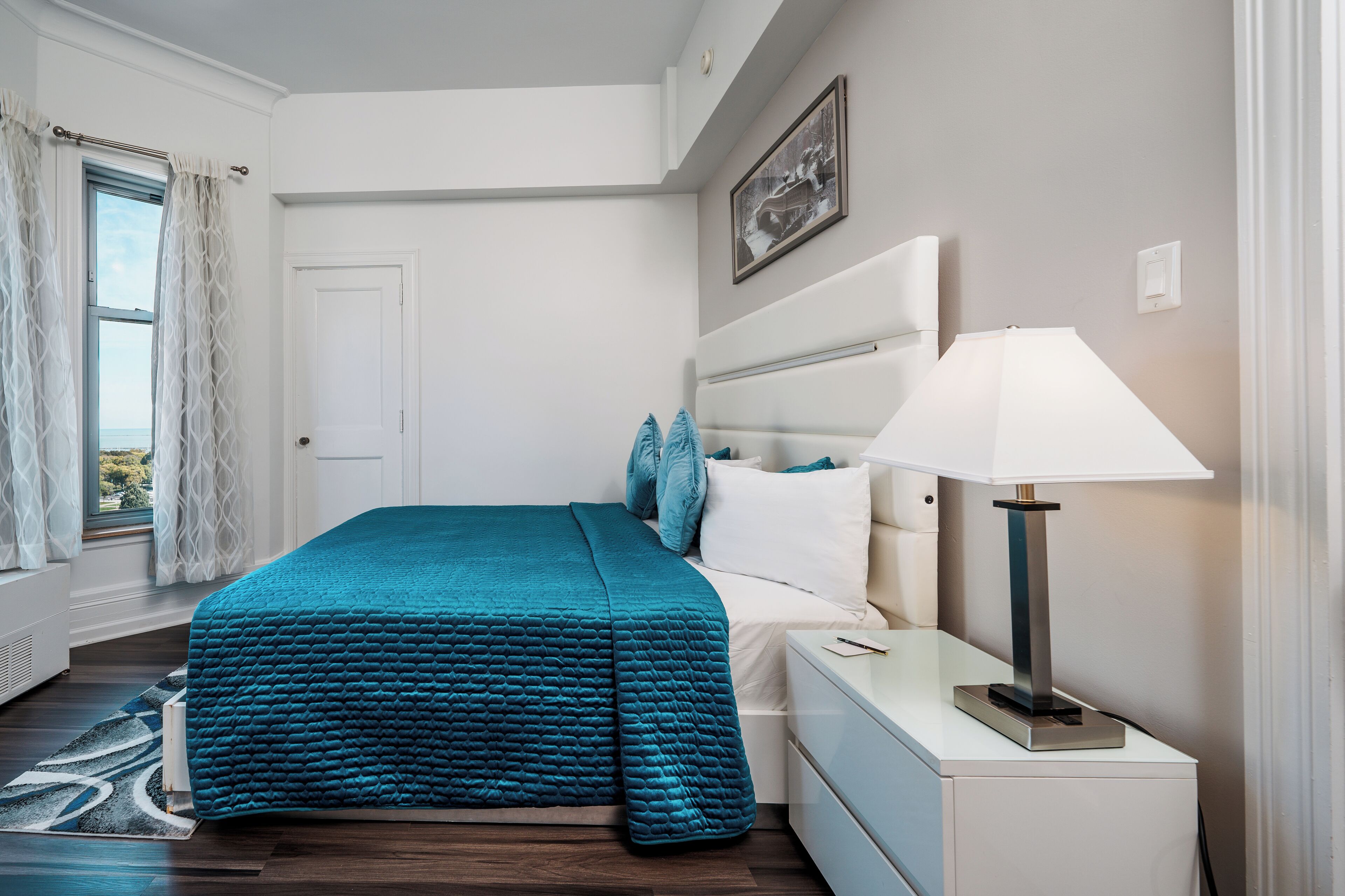 A bedroom with a bed, a nightstand, a lamp, a window with curtains, and a framed picture on the wall.