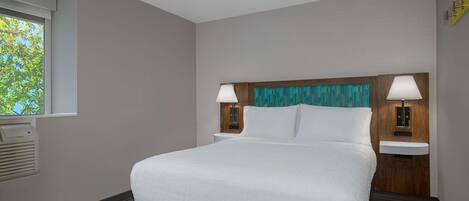 Una habitación de hotel con una cama grande, dos mesitas de noche, un ventanal con persianas, un aire acondicionado de pared y una lámpara de pared.