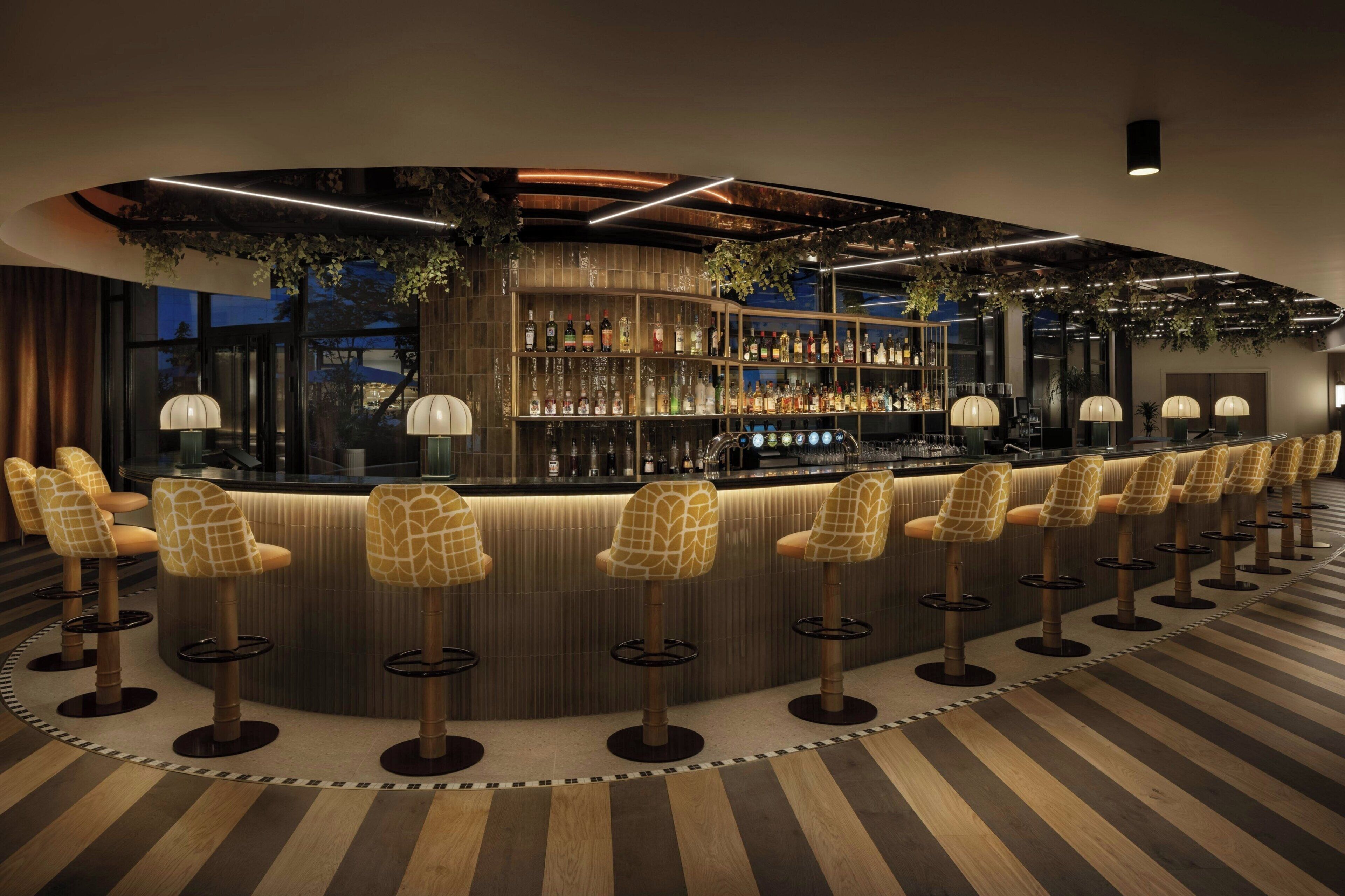 A bar area with a curved counter, multiple stools, and a variety of bottles and glasses.
