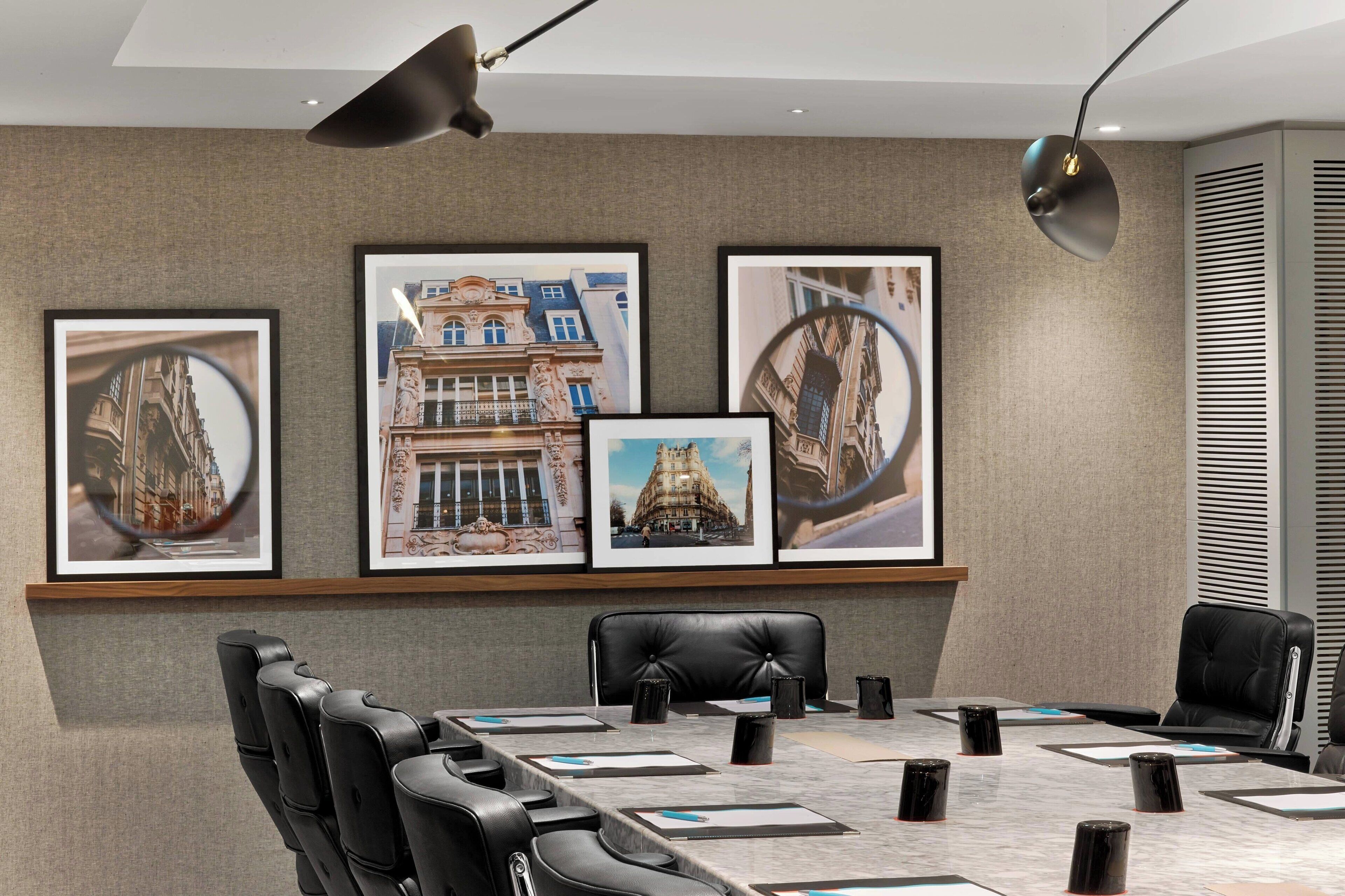 A conference room with a long table, black leather chairs, framed cityscape pictures on the wall, and a modern pendant light.