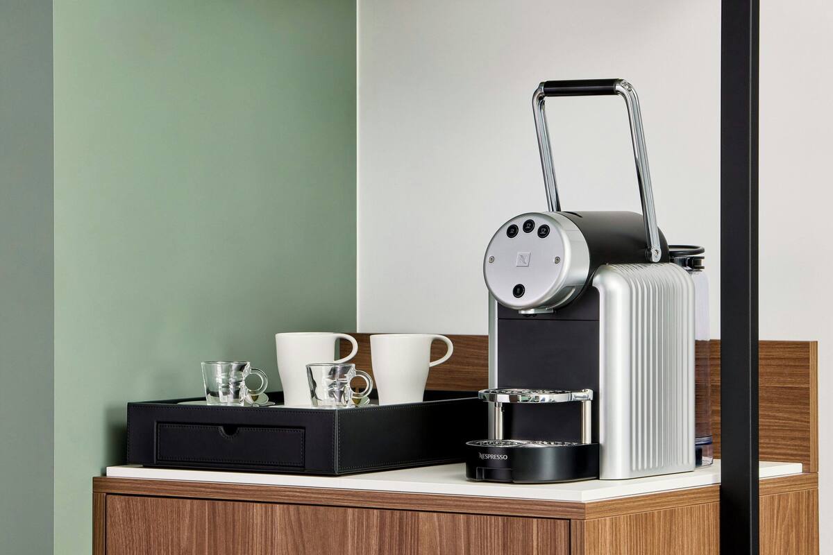 A compact espresso machine with a pull-out handle on a wooden table, accompanied by a black tray with two white cups and a glass.
