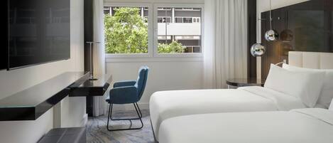 Habitación de hotel con cama, escritorio, silla, televisor y ventana con vista a edificios y árboles.