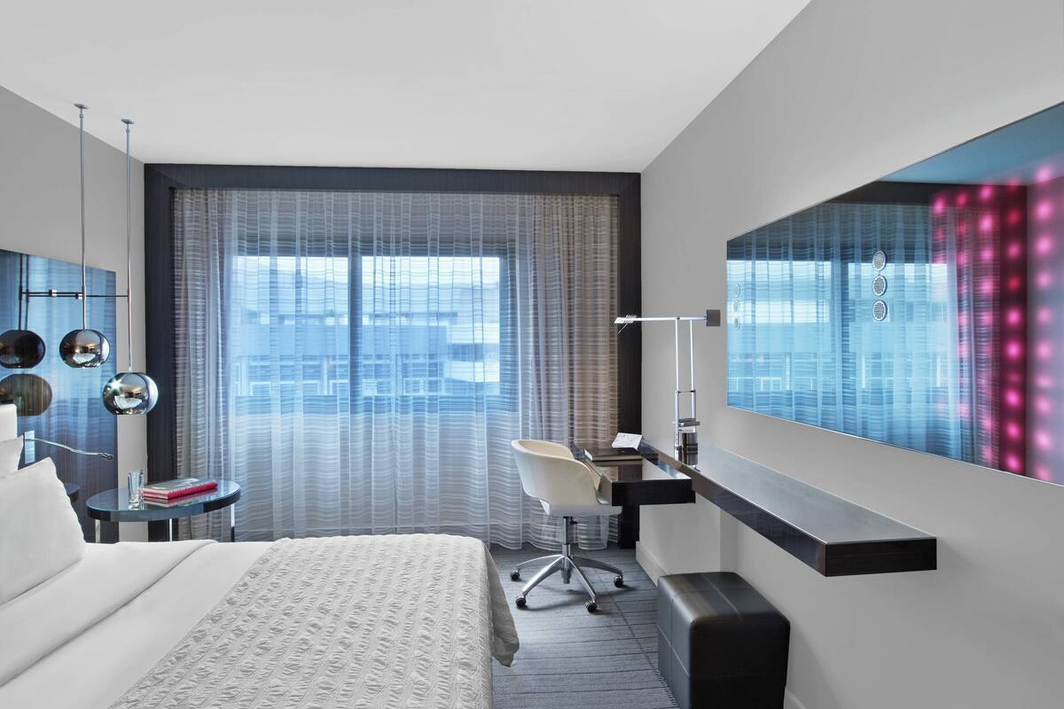 Una habitación de hotel moderna con una cama grande, un escritorio y una silla. Cuenta con un ventanal con cortinas, un televisor de pantalla plana en la pared y una mesita de noche con lámpara.