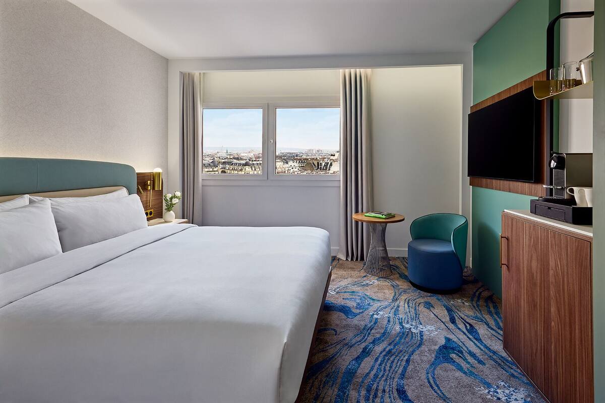 Habitación de hotel con una cama grande, vista a la ciudad, una mesita pequeña, una silla azul y un televisor.