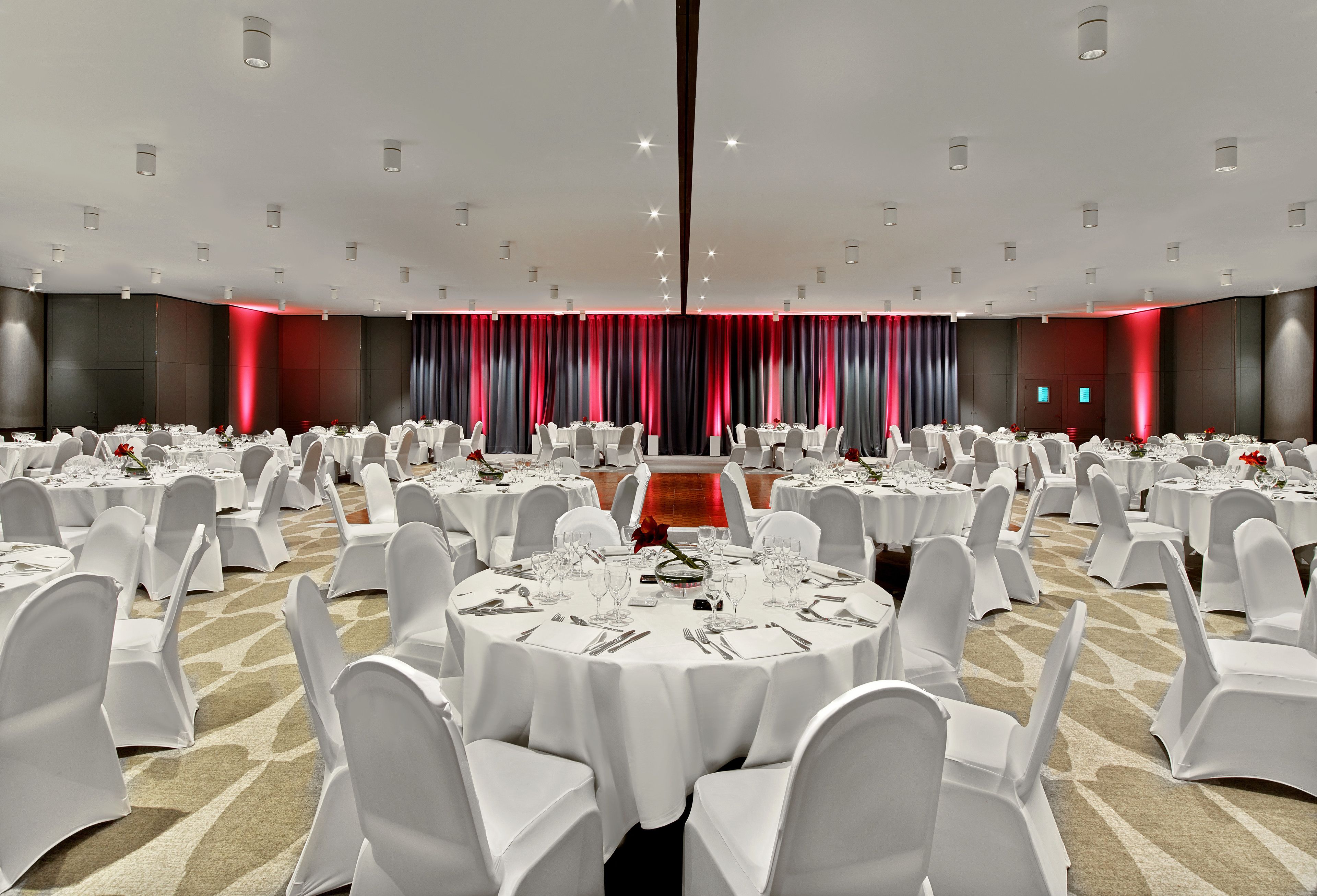 Une grande salle de banquet avec des tables rondes dressées pour le repas, des chaises blanches et un rideau rouge en arrière-plan.