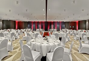 A large banquet hall with round tables set for dining, white chairs, and a red curtain backdrop.