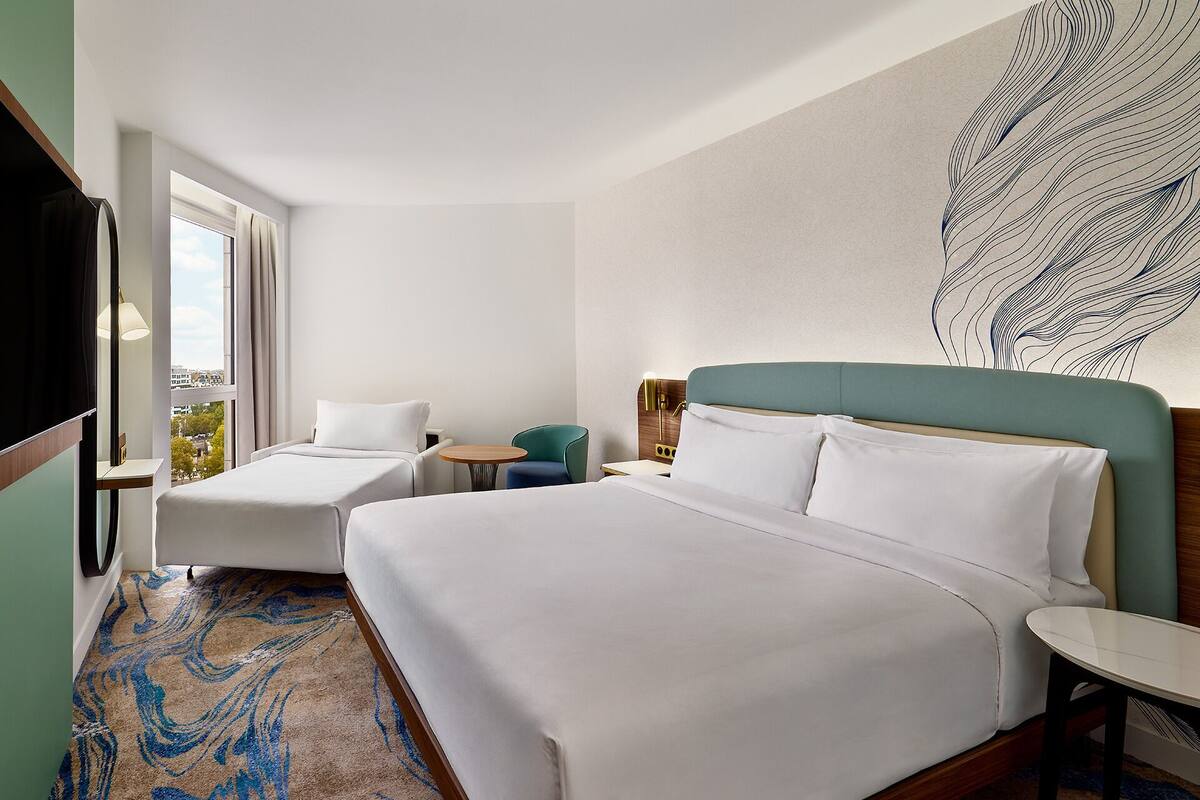 Habitación de hotel con dos camas, un escritorio, una silla y un ventanal grande.