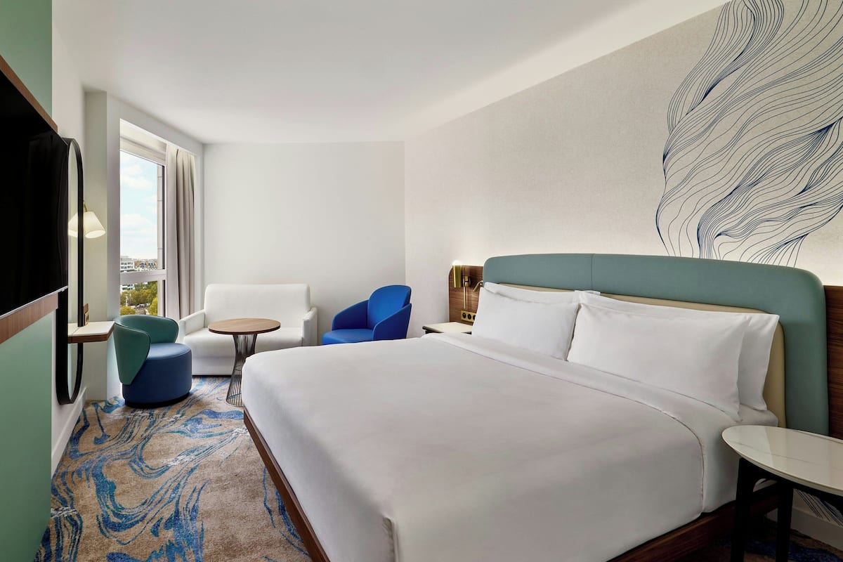 Habitación de hotel con una cama grande, un sillón azul, un sillón blanco, una mesita redonda pequeña y una obra de arte abstracta de gran tamaño en la pared.