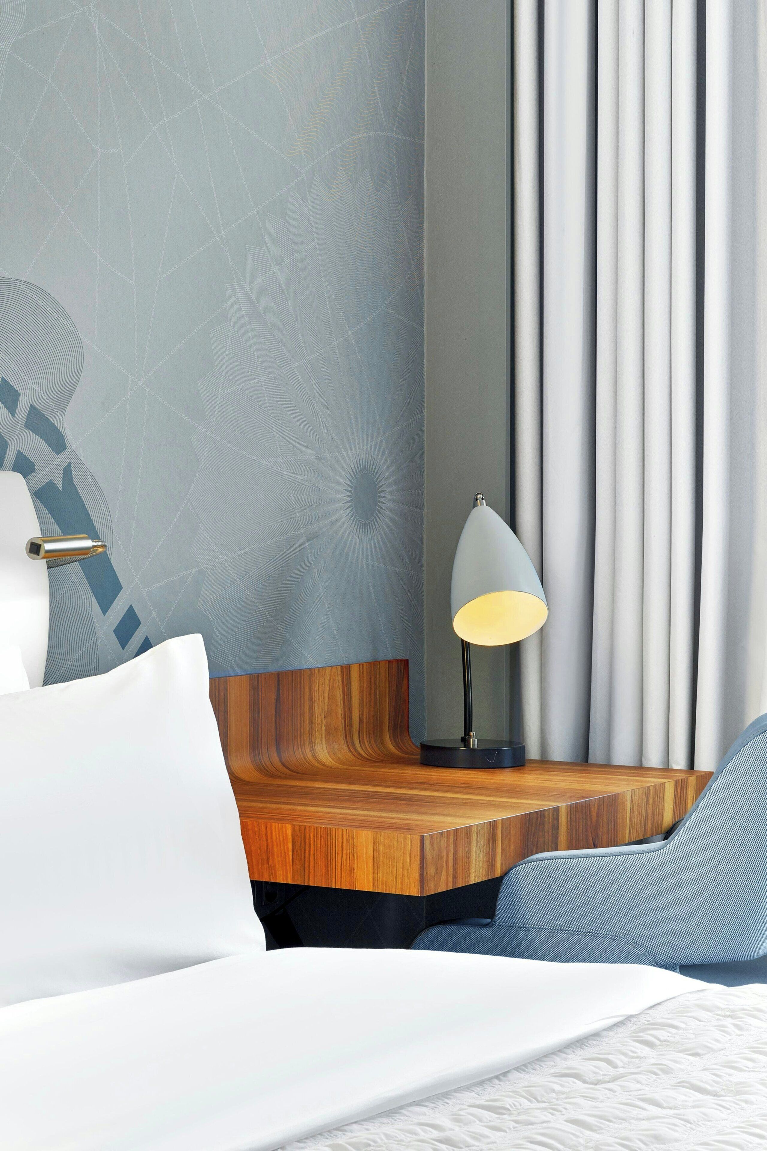 A hotel room with a bed, a nightstand, a lamp, a chair, and a curtain.