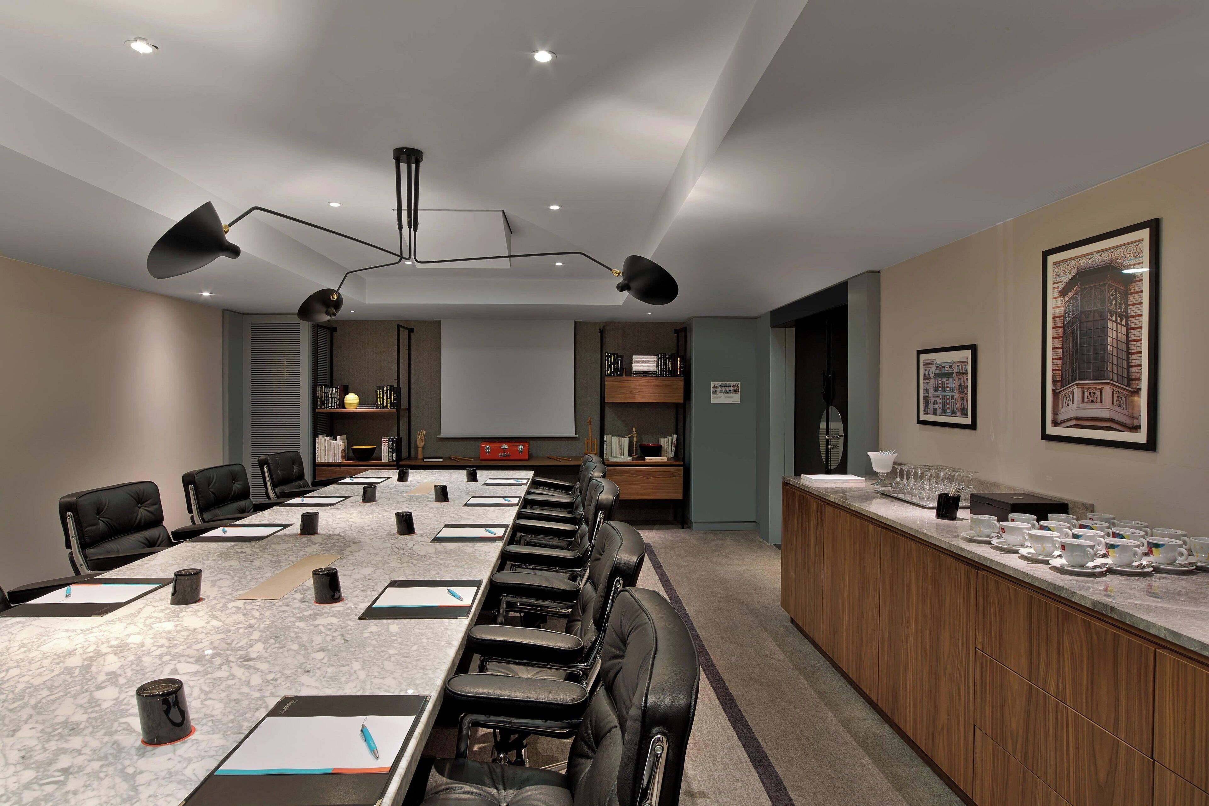 A conference room with a long table, black leather chairs, a wooden counter with cups and saucers, and framed pictures on the wall.
