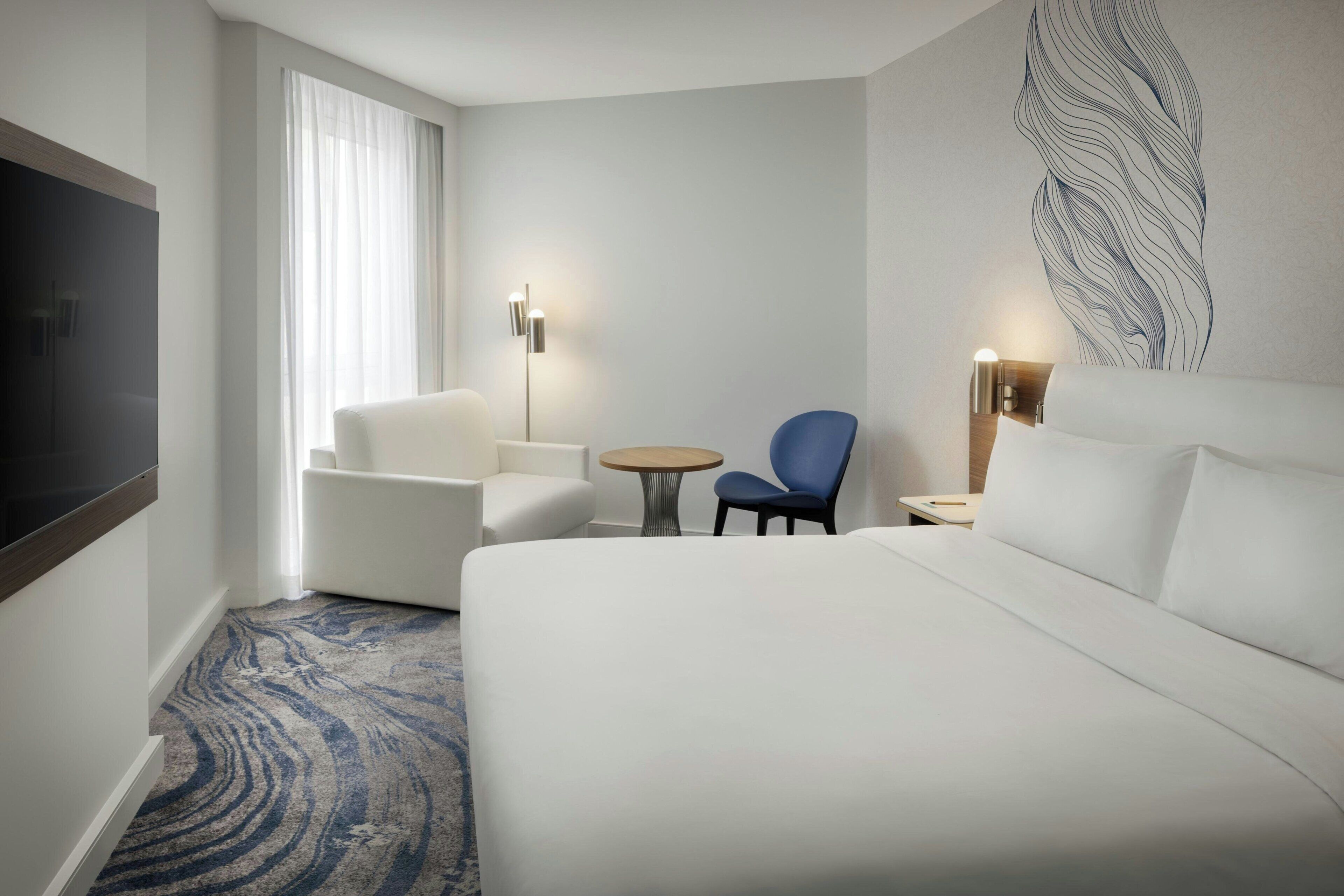 Habitación de hotel con una cama grande, una zona de estar con una silla y una mesita pequeña, un televisor de pantalla plana montado en la pared y un adorno decorativo en la pared.