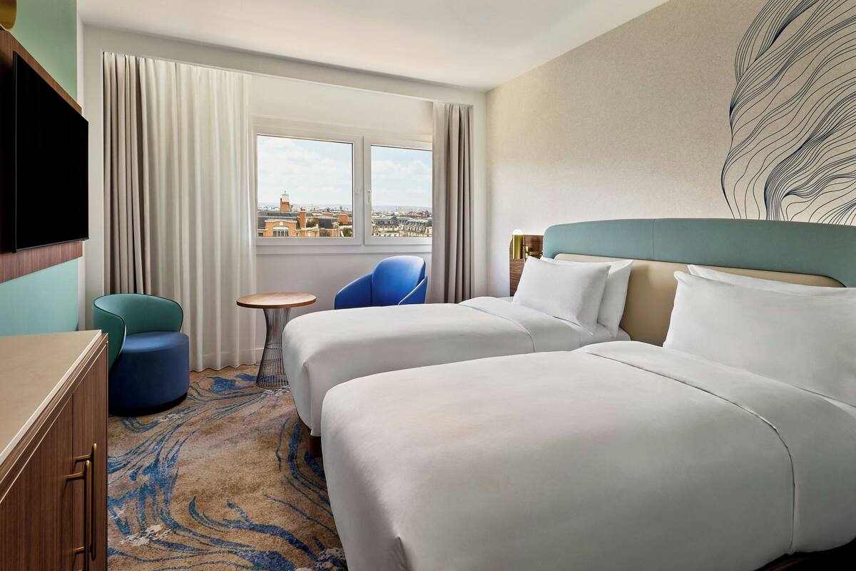 Habitación de hotel con dos camas, televisión, una mesita redonda, una silla azul y vista a la ciudad desde la ventana.