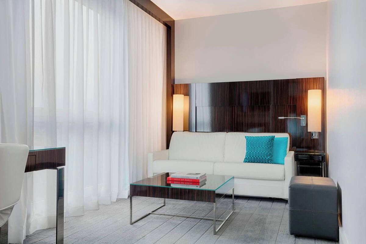 Una habitación de hotel moderna con un sofá blanco, una mesa de centro de vidrio, un taburete negro y una mesita de noche con una lámpara.