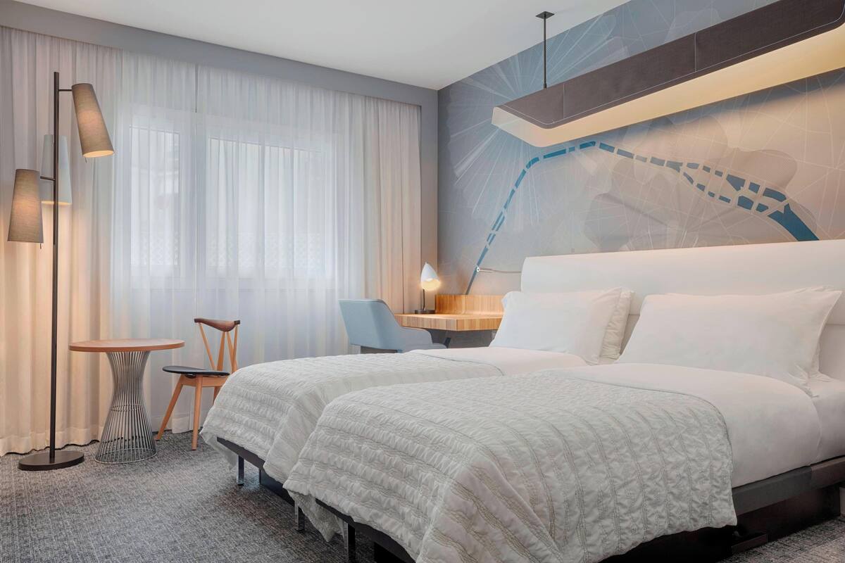 Habitación de hotel con dos camas, un escritorio, una silla, una lámpara y un ventanal con cortinas.
