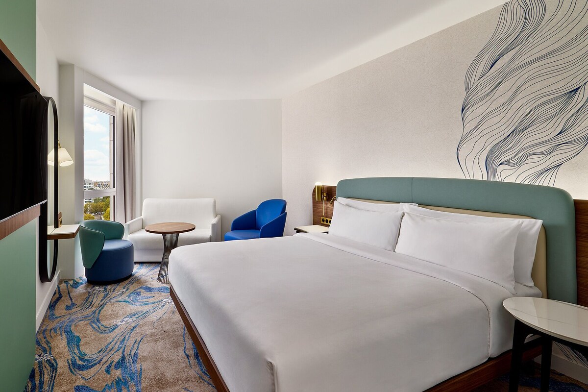 A hotel room with a large bed, a desk, a chair, and a view of the city through the window.