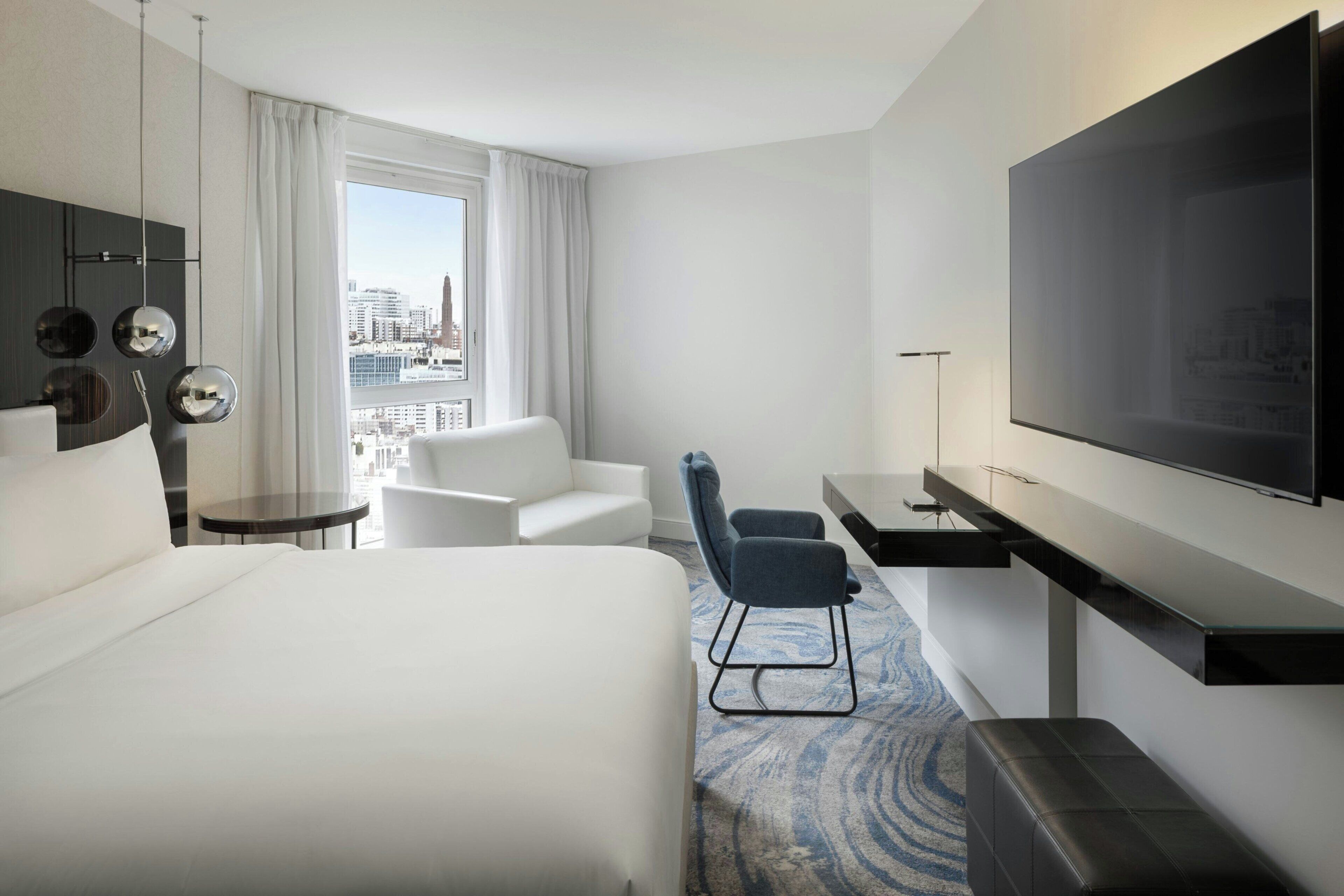 Quarto de hotel com uma cama grande, uma escrivaninha com televisão de tela plana, um espaço de estar com uma poltrona e vista para a cidade pela janela.
