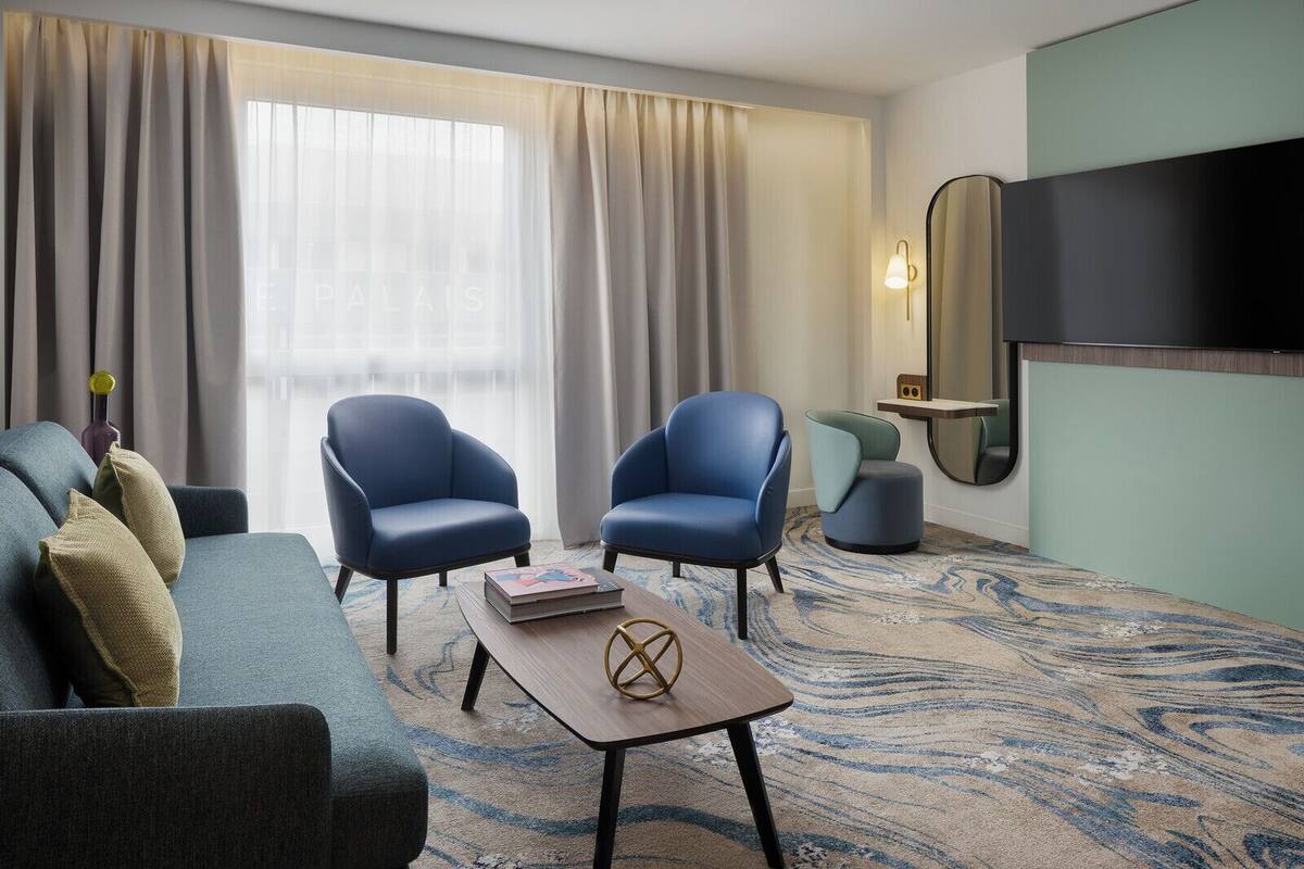 Habitación de hotel con sofá, dos sillones, una mesita de centro, televisión, espejo y alfombra estampada.
