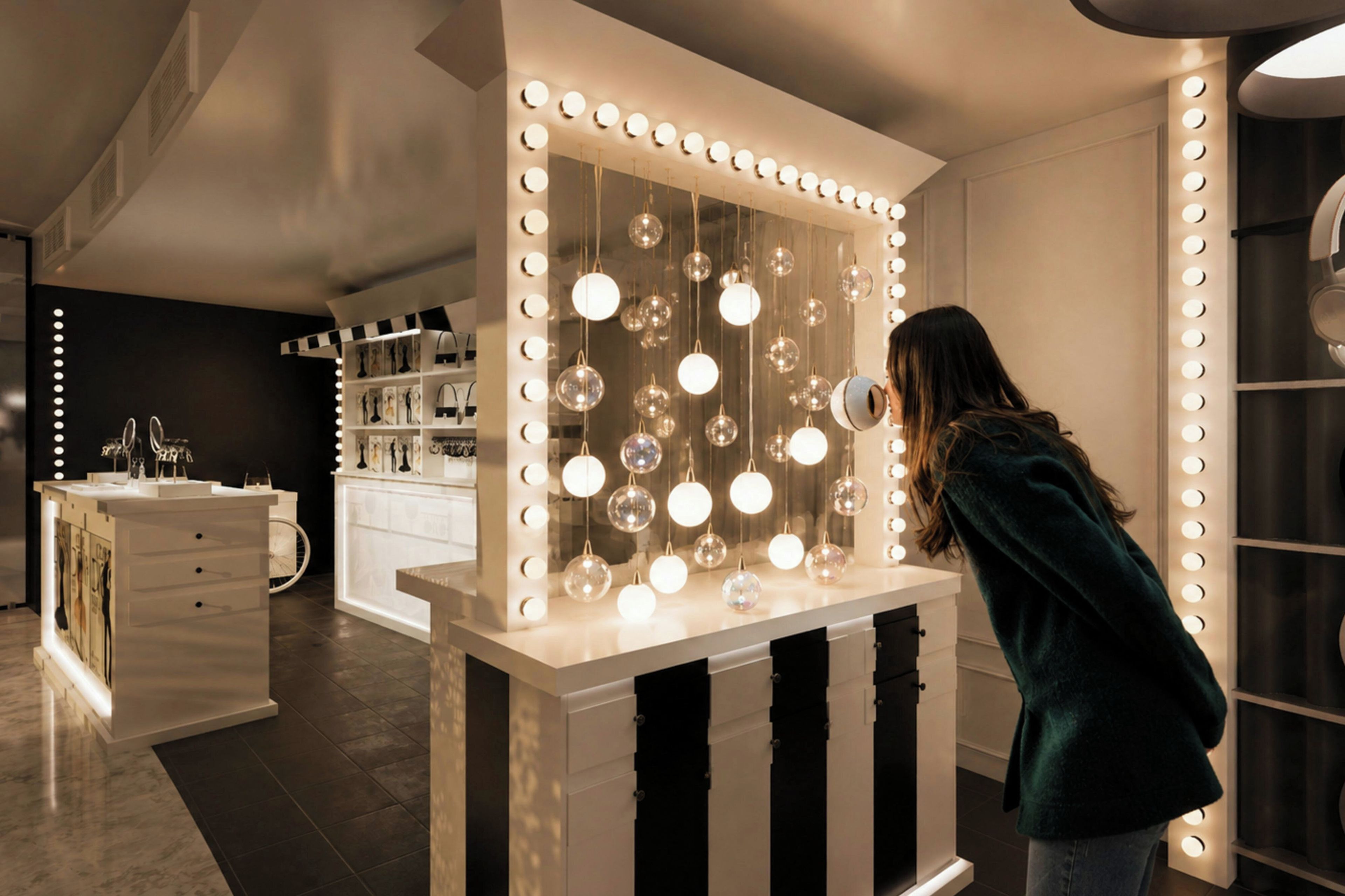A room with a vanity mirror surrounded by lights, a white dresser, and a shelf with decorative items.