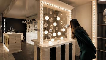 A room with a vanity mirror surrounded by lights, a white dresser, and a shelf with decorative items.