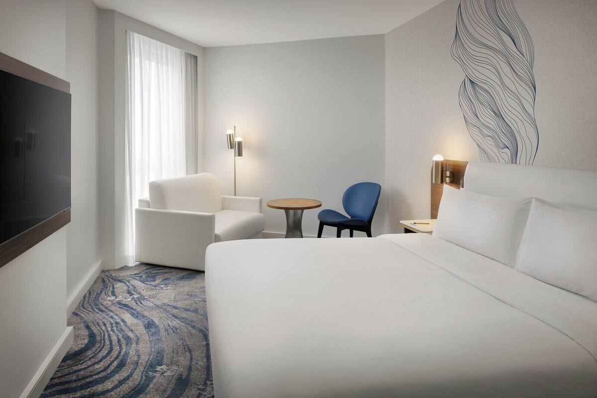 Habitación de hotel con una cama grande, un sillón blanco, una silla azul, una mesita redonda y un televisor.
