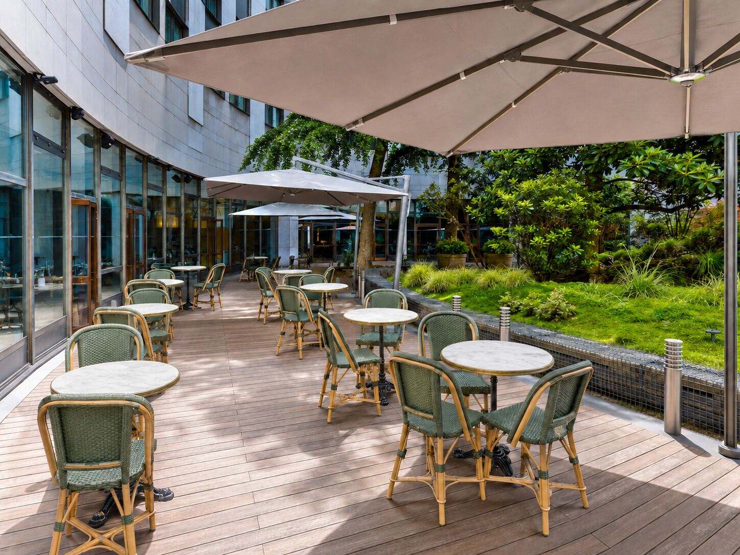 An outdoor patio with round tables and wicker chairs, surrounded by glass walls and greenery.