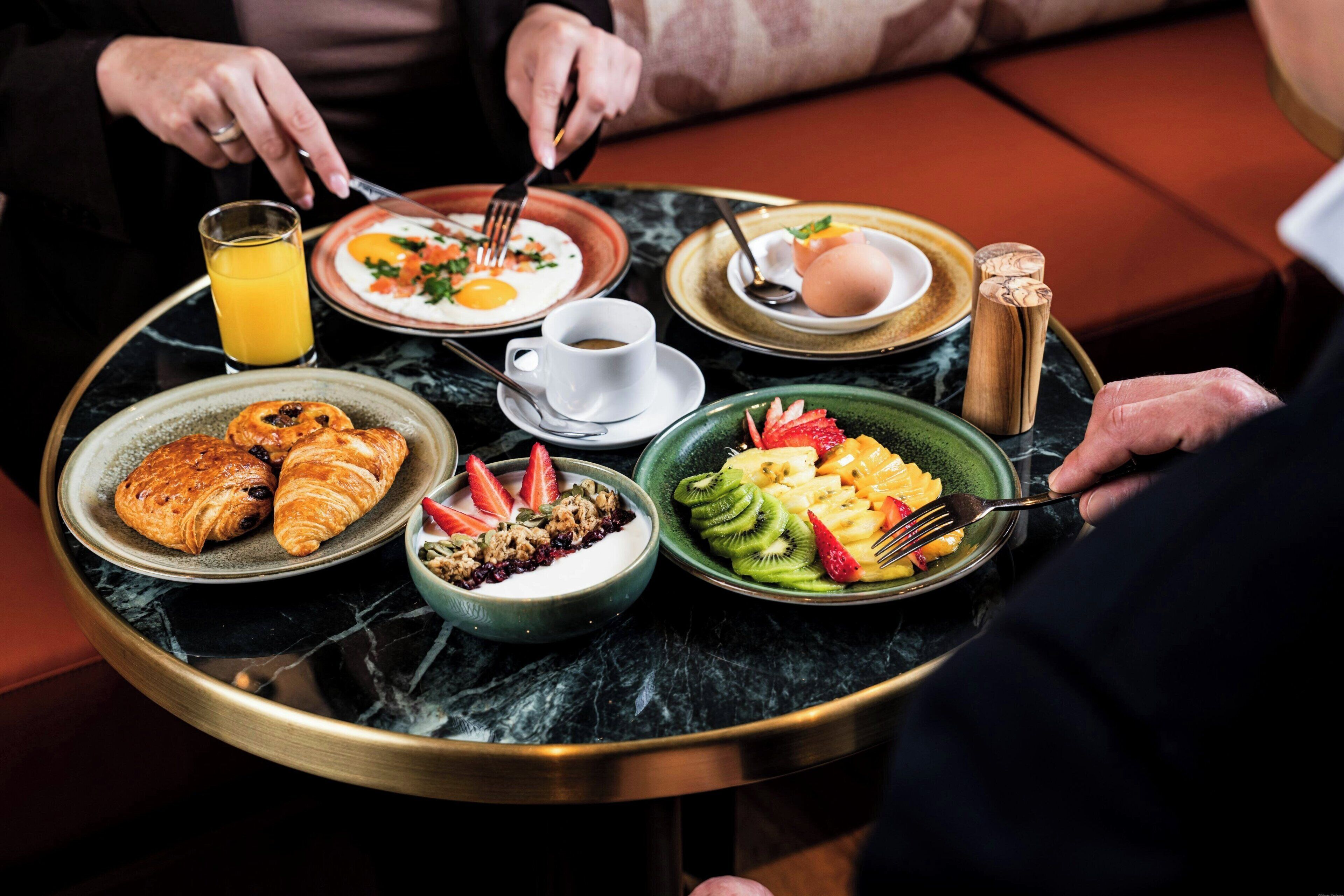 Bandeja de café da manhã com croissants, iogurte, frutas, ovos mexidos e uma xícara de café.