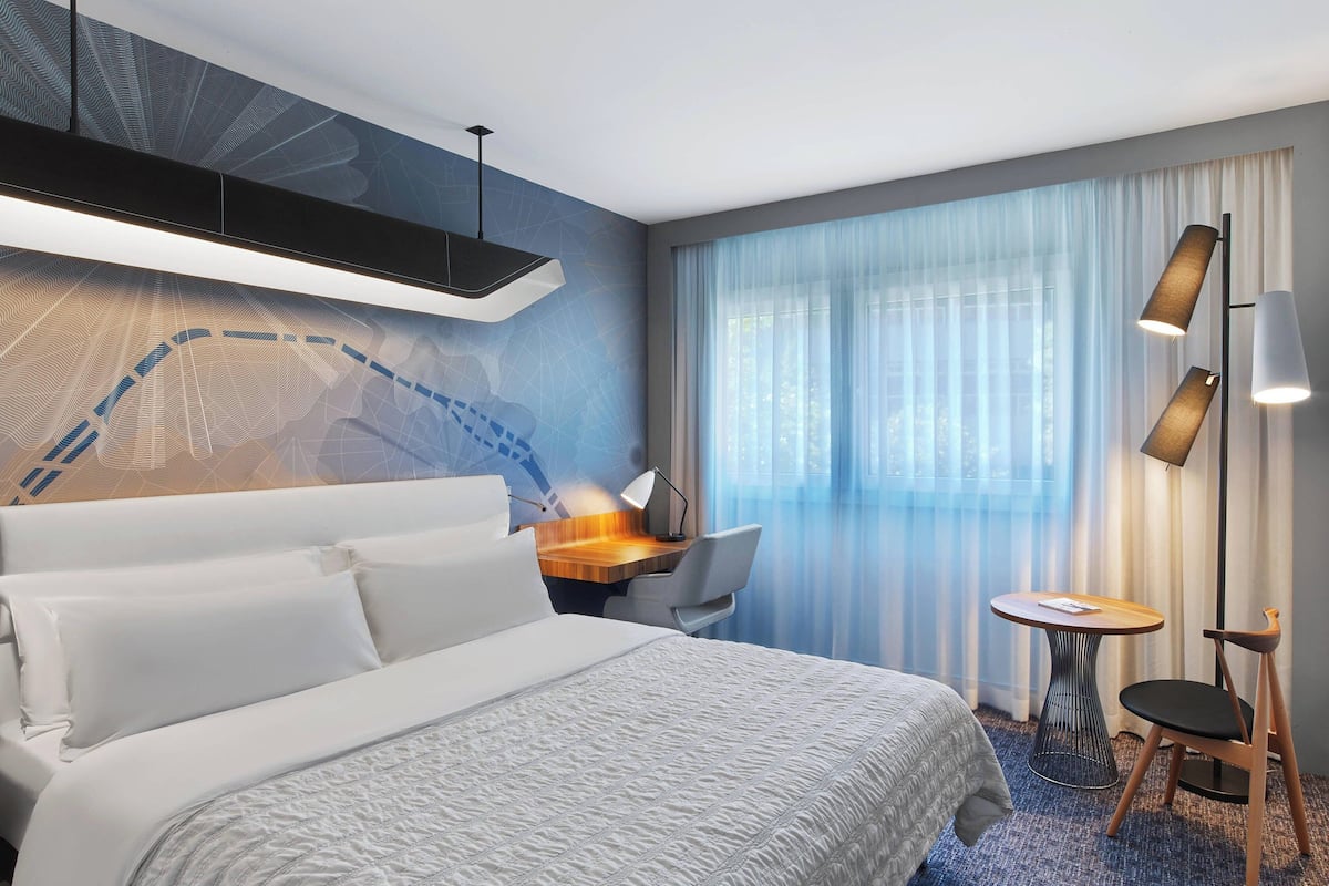 A modern hotel room with a large bed, a desk, a chair, a small table, and a floor lamp.