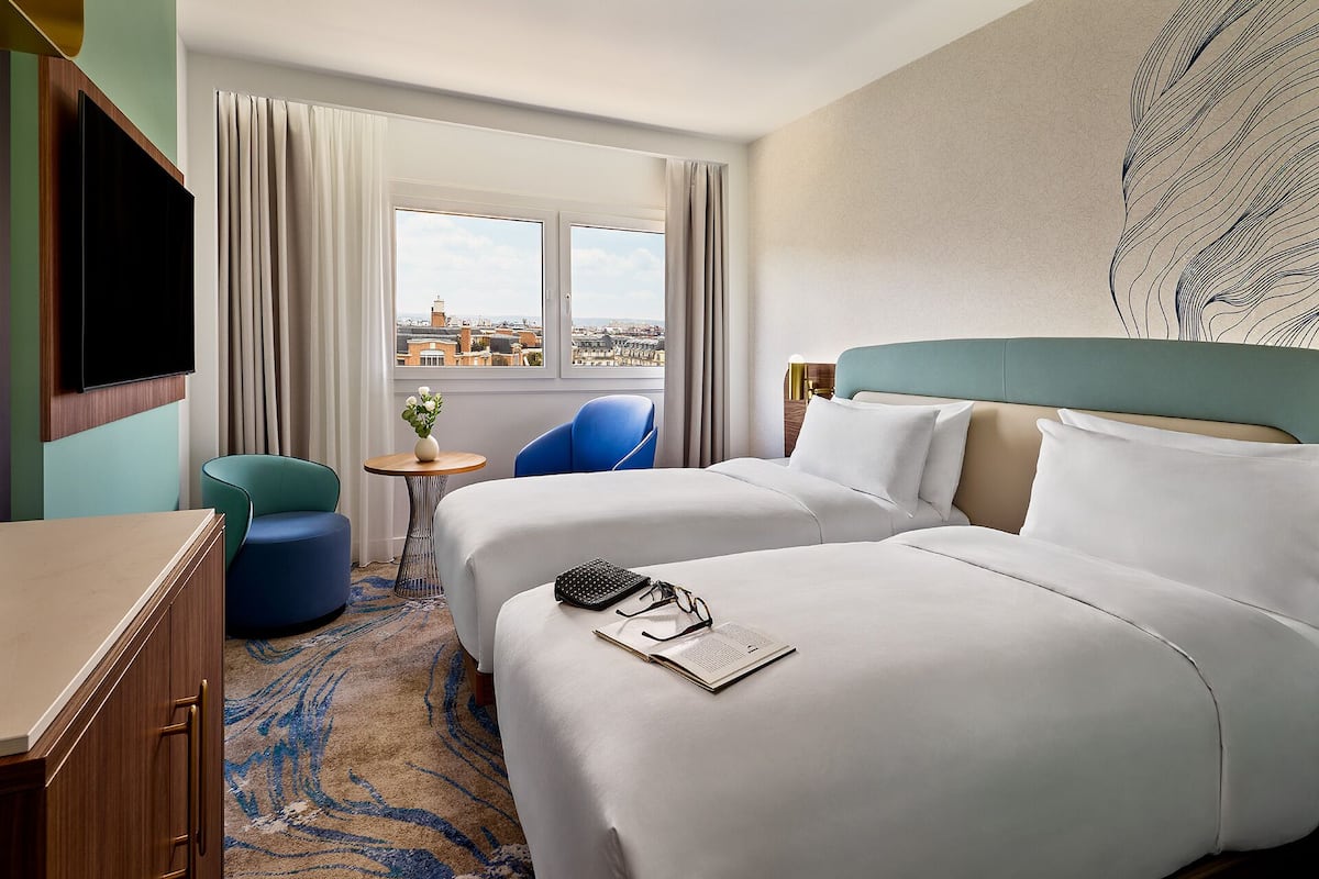 A hotel room with two beds, a desk, a chair, a small table with a vase, and a city view through the window.