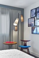 Ett hotellrum med en säng, ett litet runt bord med en bok, en blå stol, en golvlampa och inramade tavlor på väggen.