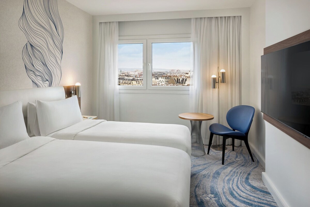 A hotel room with two beds, a TV, a small table, a chair, and a view of the city.
