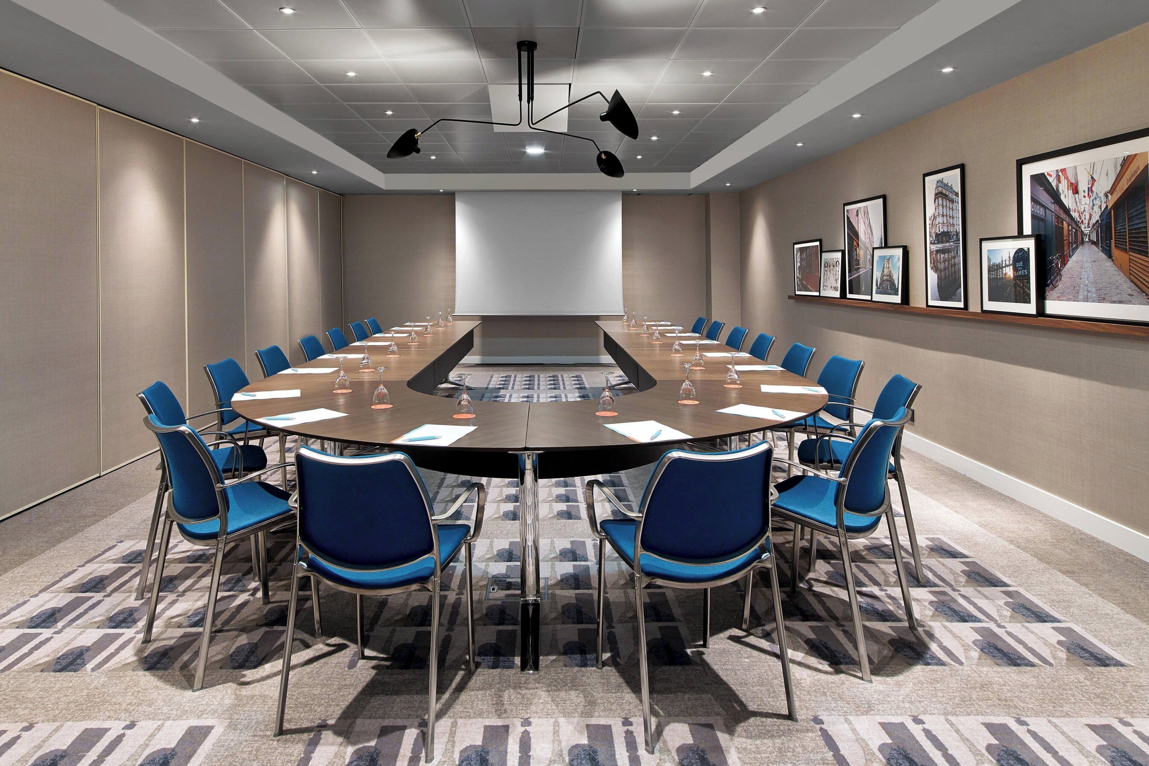 A conference room with a large oval table, blue chairs, a projector screen, and framed pictures on the wall.