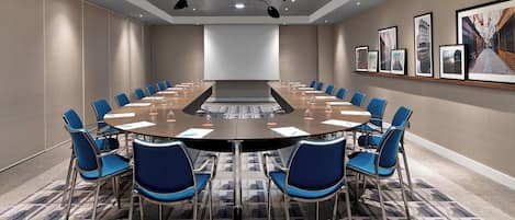 A conference room with a large oval table, blue chairs, a projector screen, and framed pictures on the wall.