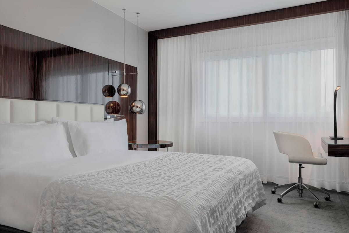 A modern hotel room with a large bed, a desk, and a chair. There is a large window with sheer curtains and a mirror on the wall.
