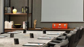 Een vergaderruimte met een lange tafel, zwarte stoelen, een scherm voor een beamer, een boekenplank en een kleine oranje koffer.
