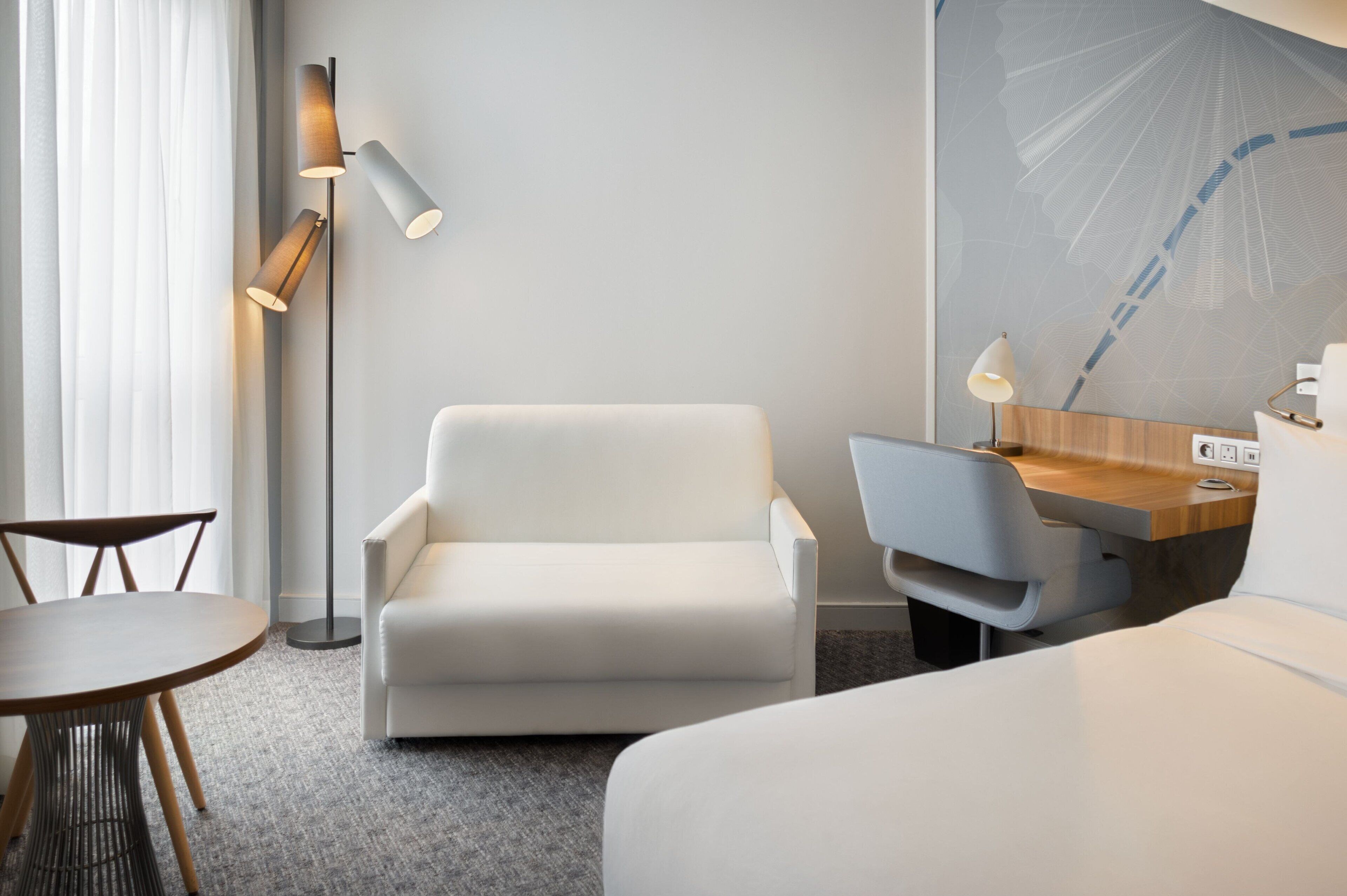 Una habitación de hotel moderna con un sillón blanco, un escritorio con lámpara, una mesa redonda y un gran mural abstracto en la pared.