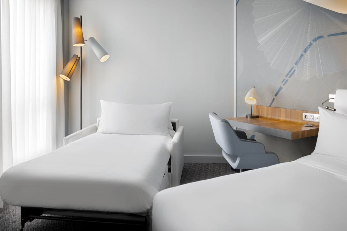 A hotel room with two beds, a desk, a chair, a lamp, and a wall-mounted light fixture.