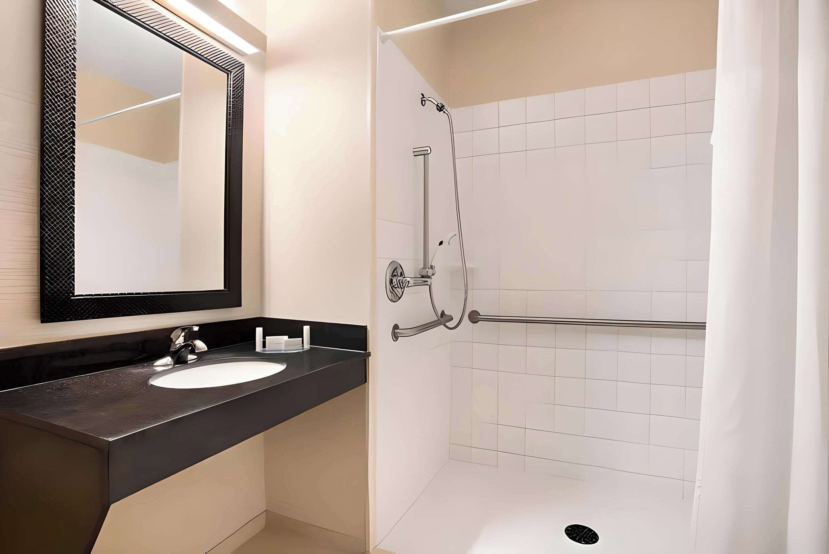 Une salle de bain avec un lavabo sur un plan de toilette noir, un grand miroir et une douche équipée d’une barre d’appui.