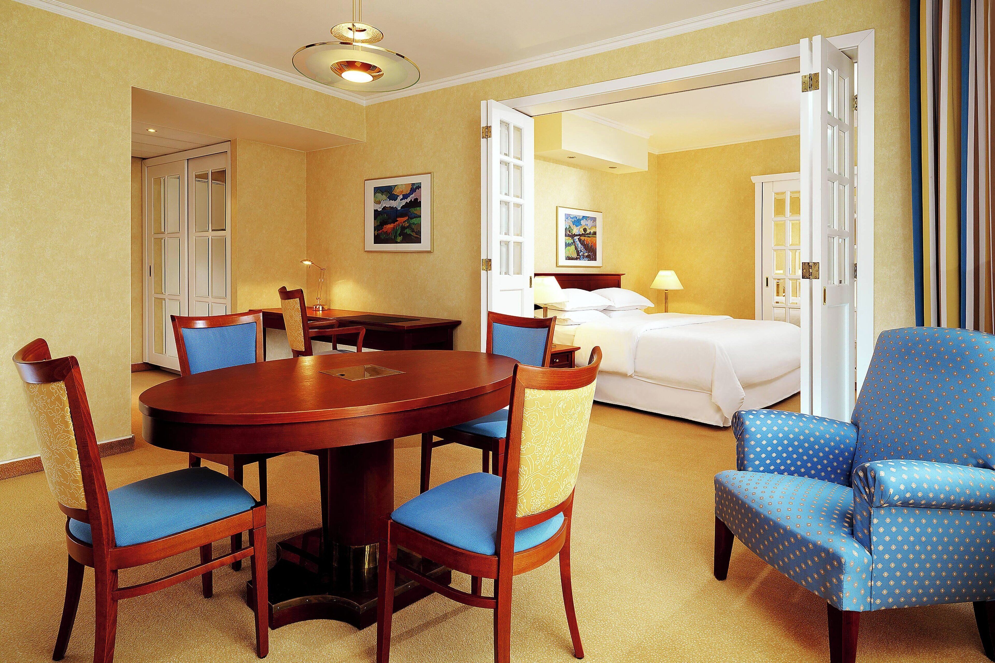 Habitación de hotel con una mesa de comedor, sillas, una cama y un baño visible a través de una puerta abierta.