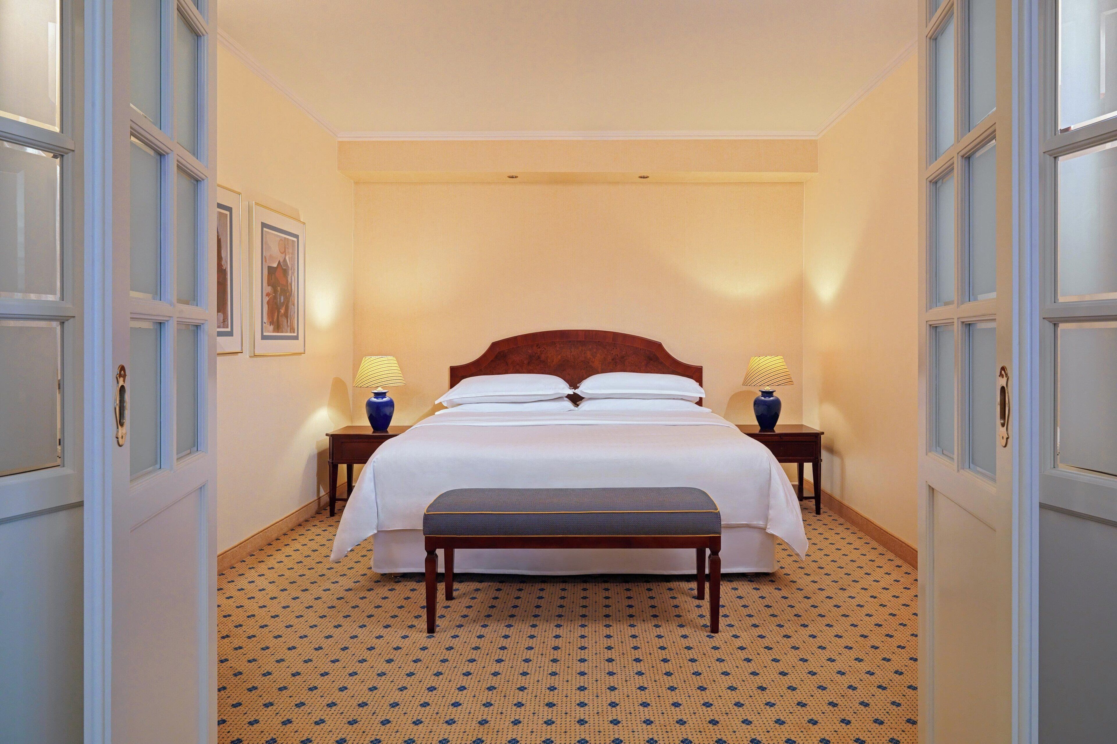 Een hotelkamer met een groot bed, twee nachtkastjes met lampen, een bankje en een kamerbreed tapijt met dessin.