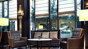 A modern hotel lobby with a sofa, two armchairs, a round table, and large windows with a view of trees. - Sheraton Essen Hotel (Essen)