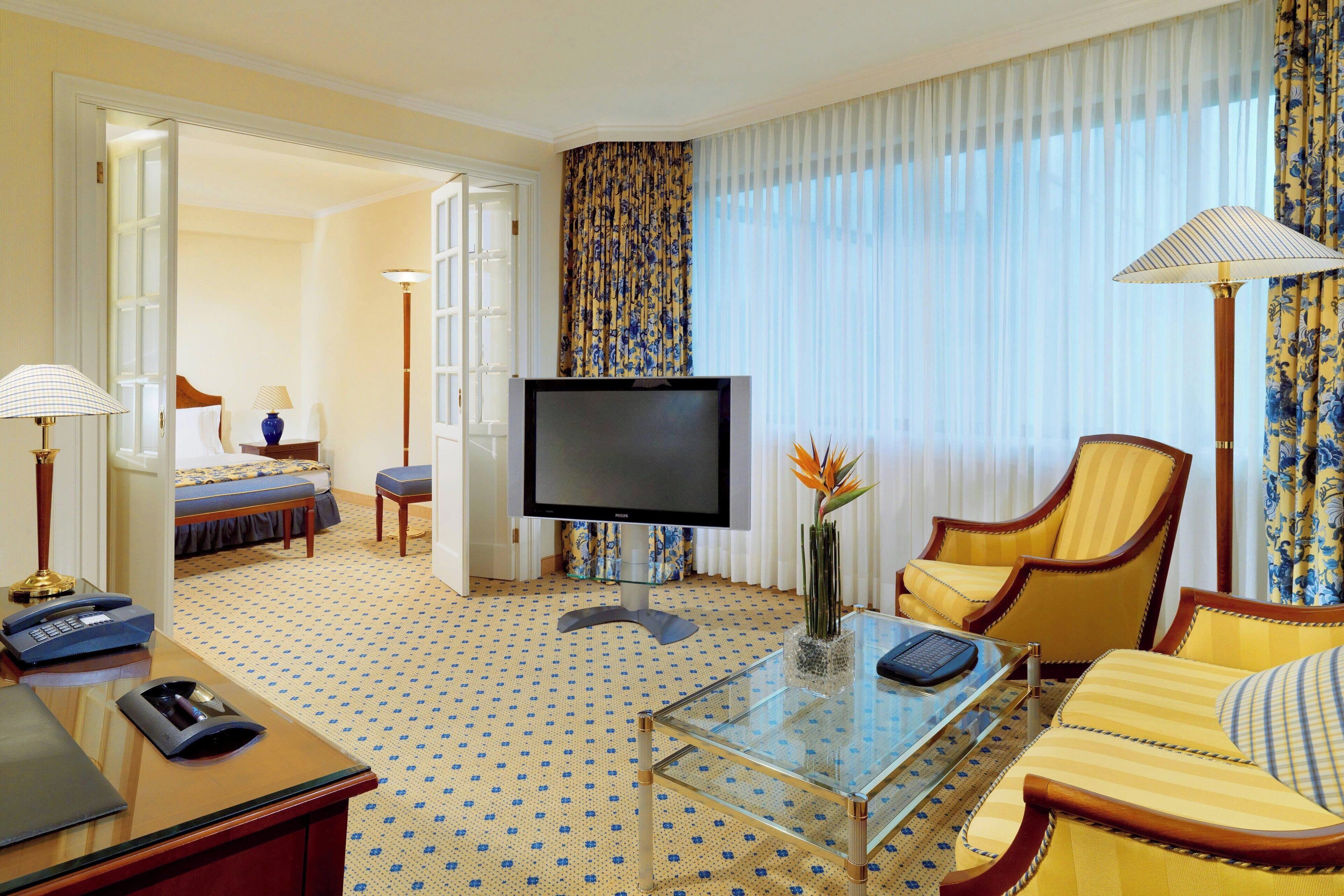 A hotel room with a television, a glass coffee table, a sofa, a chair, a desk with a telephone, and a bed with a blue and white patterned bedspread.