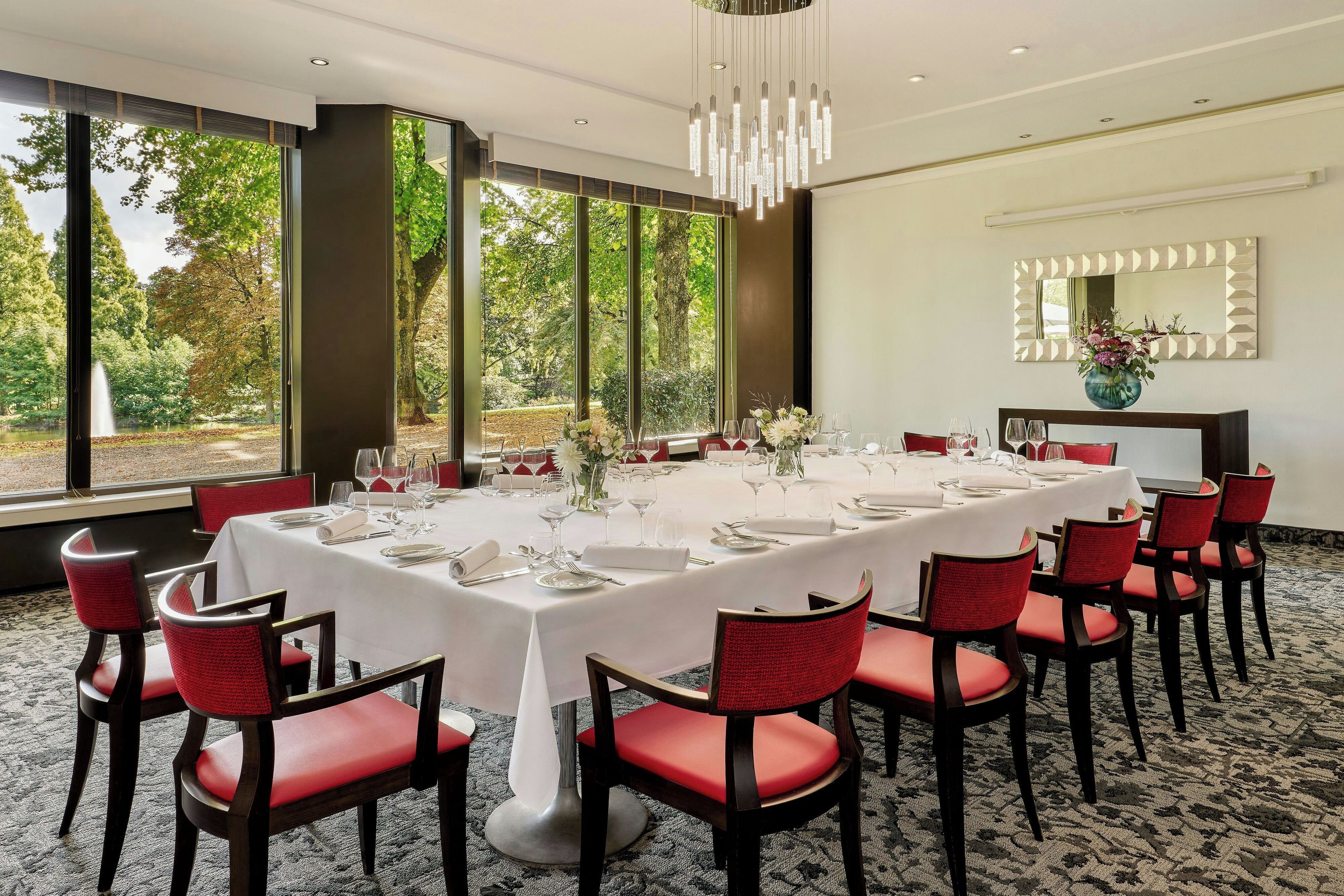 Un coin repas avec une grande table dressée pour un repas, entourée de chaises rouges, et offrant une vue sur des arbres et une fontaine à l’extérieur.