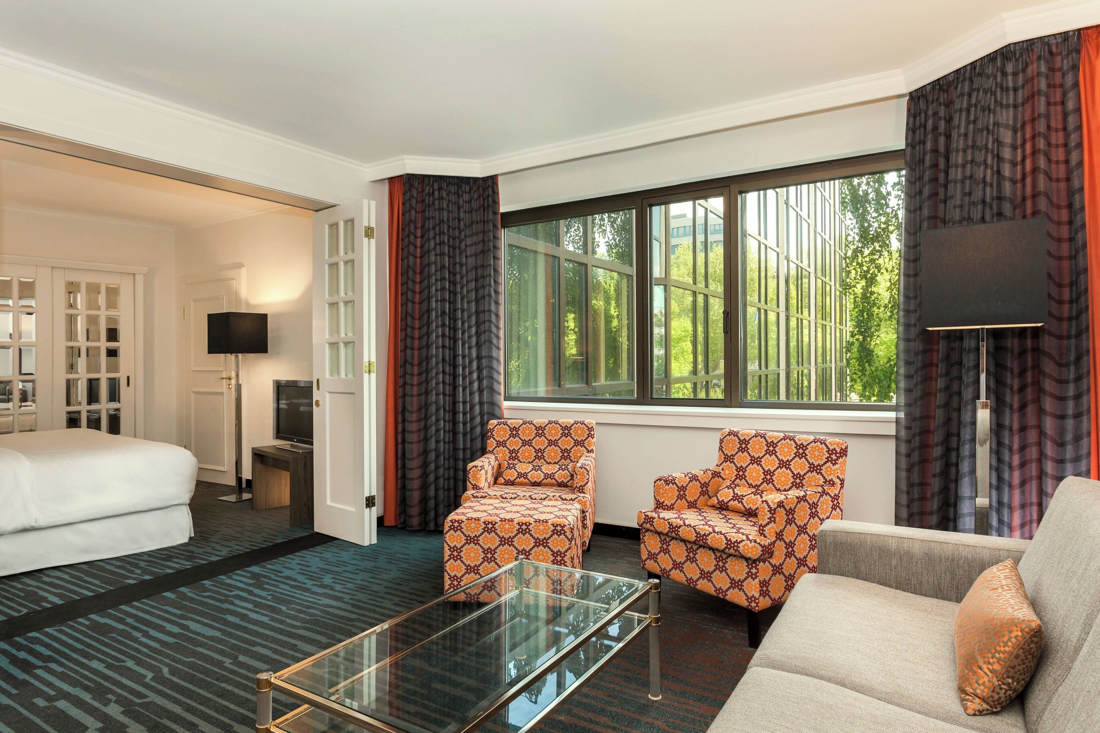A hotel room with a bed, two patterned armchairs, a sofa, a glass coffee table, a TV, and a large window with curtains.