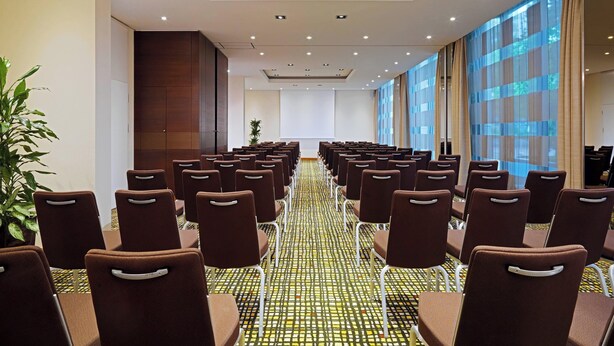 A conference room with rows of brown chairs, a patterned carpet, and large windows with curtains. - Sheraton Essen Hotel (Essen)