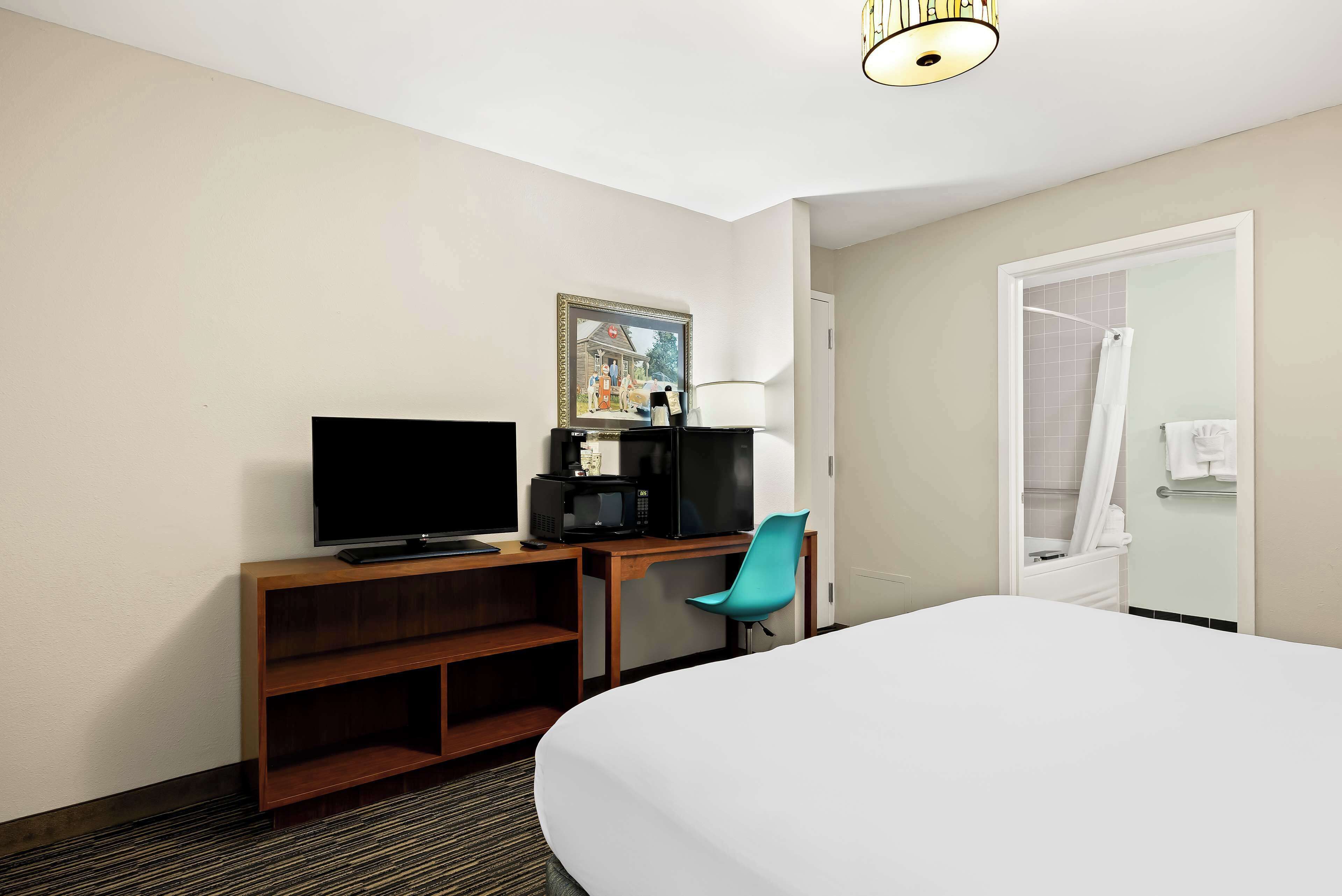 Habitación de hotel con cama, escritorio con televisor, silla, microondas y un cuadro en la pared.