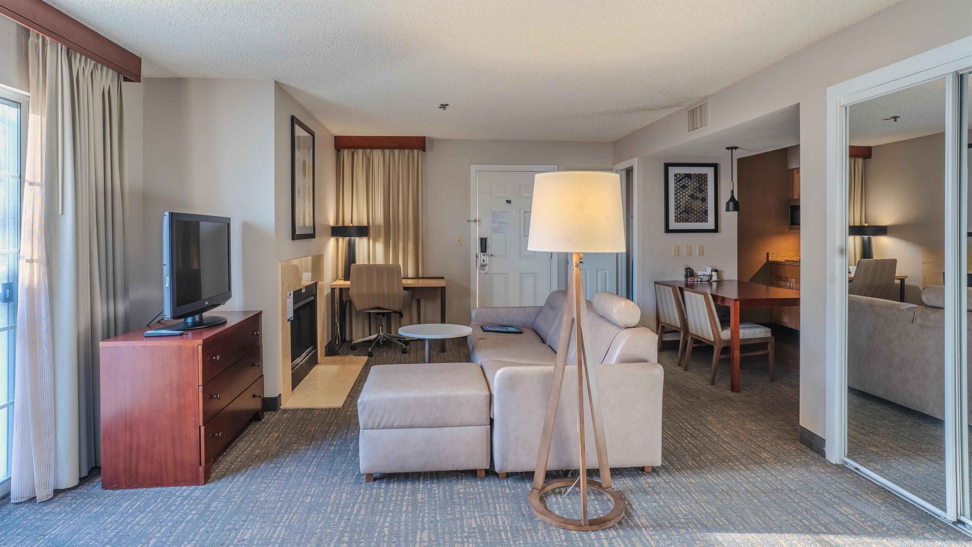 A hotel room with a television, sofa, armchair, dining table, and a lamp.